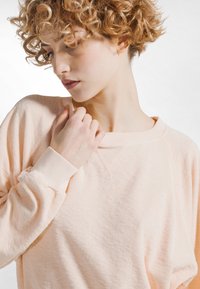 Vaalean persikanvärinen pitkähihaisen collegepaita rentolla istuvuudella, resorit hihoissa ja pehmeä, teksturoitu kangas. Siinä on leveä pääntie ja hienovaraiset saumat.