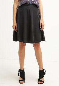 Jupe noire en forme A en tissu lisse, arrivant aux genoux avec une taille ajustée et un ourlet évasé, associée à des bottes à cheville noires ouvertes.