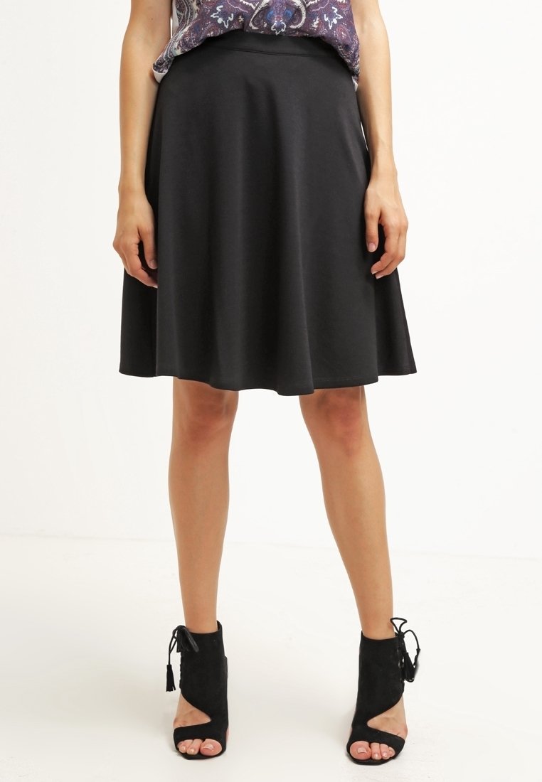 Jupe noire en forme A en tissu lisse, arrivant aux genoux avec une taille ajustée et un ourlet évasé, associée à des bottes à cheville noires ouvertes.