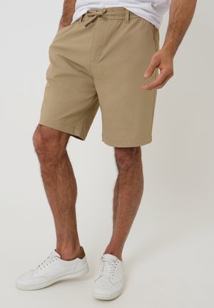 Mand iført beige knælange shorts med snøre i taljen og hvide sneakers, stående foran en ensfarvet lys baggrund.