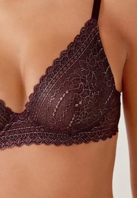 Bralette di pizzo in un profondo bordeaux con motivi floreali, bordi ondulati e spalline regolabili su un tono di pelle chiaro.
