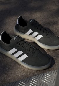 Sneakers Adidas nere con strisce bianche e suole traslucide poggiate su una superficie texturizzata di cemento alla luce naturale.