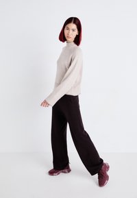Pull en maille beige clair avec col montant et ourlet côtelé, associé à un pantalon large sombre et des chaussures de sport bordeaux.