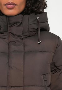 pruun puffer jope kõrge kraega, kokku tõmmatava kapuutsiga, horisontaalse õmbluse disainiga ja külgmistest lukuga taskutest. Lihtne, mati tekstuuriga.