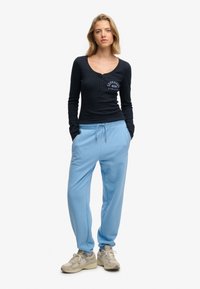 Haut henley à manches longues bleu marine avec détails à boutons et pantalon de survêtement bleu. Le matériau est doux, avec une coupe décontractée. Baskets crème avec accents texturés.