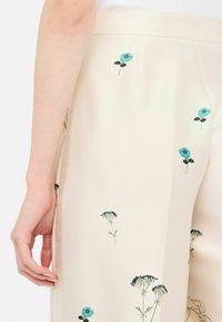 Pantaloni crema con ricami floreali in tonalità di blu e verde, caratterizzati da una texture liscia e un design a gamba larga.