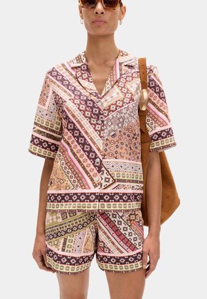 Kvinde iført mønstret kortærmet skjorte med knapper og matchende shorts, bærende en brun ruskindstaske, iført store solbriller.