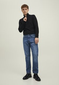 Svart ribbestrikket genser med høy krage, kombinert med lyseblå rettlagde jeans og svarte snøresko. Nøytral bakgrunn.