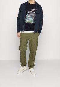 Giacca zip navy sopra una t-shirt nera con grafiche, abbinata a pantaloni cargo verde oliva e sneaker chiare. La maglietta presenta un design naturalistico.