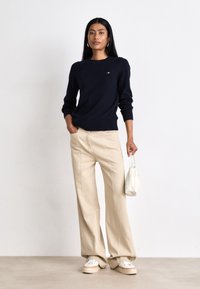 Maglione blu navy a maglia con collo e polsini a coste, abbinato a pantaloni beige larghi. Gli accessori includono una piccola borsa bianca e sneakers casual.