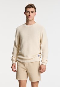 Beige knitted sweater with a textured pattern, round neckline, and ribbed cuffs; features a small blue emblem at the hem.