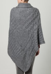 Grå stickad poncho med ett komplext mönster som har flätor och texturerade stygn, avslutad med en bred krage och asymmetrisk fåll.