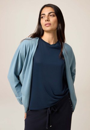 Helles blaues Cardigan mit lockerer Passform und gerippten Manschetten, getragen über einem dunkelblauen Oberteil. Glatte Textur und schlichtes Design, geeignet zum Überziehen.