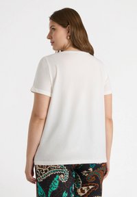 Camiseta blanca de manga corta con puños enrollados, hecha de tejido suave, con un diseño sencillo. El modelo lleva pantalones estampados en marrón y azul verdoso.