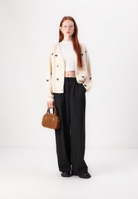 Gilet crème avec des motifs de nœuds noirs, crop top blanc, pantalon noir à large jambe et un petit sac à main en cuir texturé marron. Des chaussures noires complètent le look.