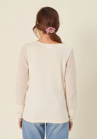 Femme aux cheveux bruns attachés avec un chouchou rose à motifs, portant un pull crème à manches longues ajourées et un jean bleu, vue de dos.