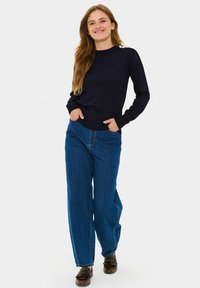 Granatowy sweter warkoczowy z ribbami na mankietach i okrągłym dekoltem, połączony z jasnoniebieskimi spodniami w szerokim kroju. Dodatki to stylowe brązowe buty.