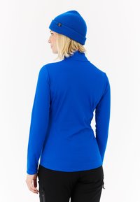 Plava majica dugih rukava s visokim ovratnikom, izrađena od glatkog, rastezljivog materijala. Nosena s crnim hlačama i skladnom plavom pletenom kapom.