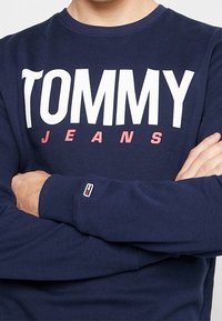 Námořnická modrá mikina s velkým bílým nápisem "TOMMY" a menším růžovým "JEANS" pod ním. Má ribbované manžety a logo značky na rukávu.
