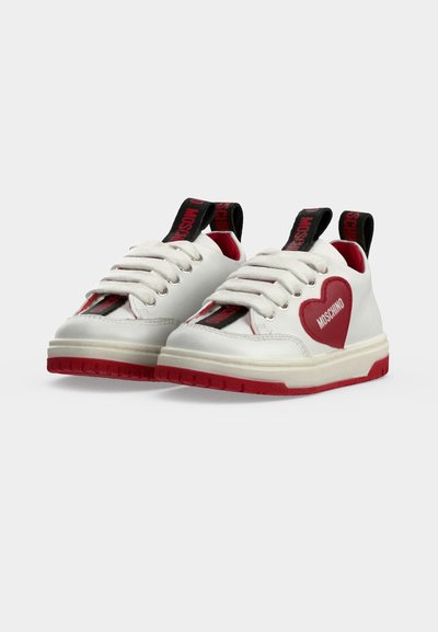Baskets blanches avec un détail cœur rouge sur les côtés, semelles rouges, languettes noires avec texte rouge et lacets blancs sur un fond uni.