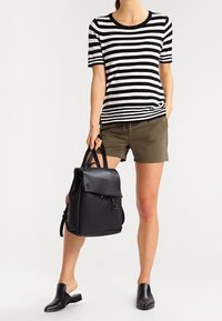 Sac à dos en cuir noir avec rabat et poche avant, associé à un t-shirt rayé noir et blanc, des shorts olive et des chaussures noires sans lacets.