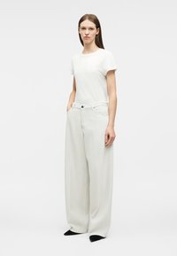 Γυναίκα στέκεται με τα χέρια στις τσέπες, φοράει λευκή μπλούζα με κοντά μανίκια, λευκό παντελόνι φαρδύ και μαύρα μυτερά παπούτσια σε απλό φόντο.