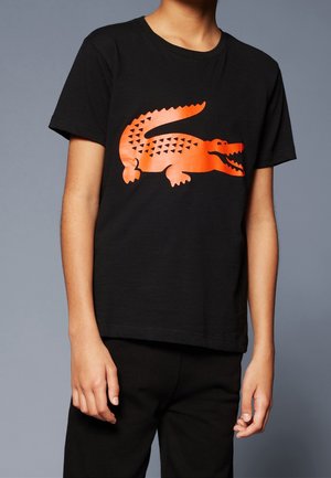 Junge Person trägt ein schwarzes Kurzarmshirt mit einem großen, leuchtend orangefarbenen Alligator-Design auf der Brust vor einem schlichten grauen Hintergrund.