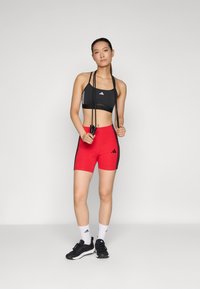 Crna sportska grudnjak s tri bijele pruge, u paru s crvenim šortsem koji ima bočne pruge i sportski dizajn. Crne tenisice i uže.
