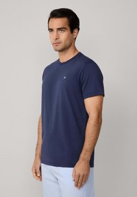Námořnická modrá bavlněná tričko s kulatým výstřihem, krátkými rukávy a malým logem na levé části hrudi. Hladká textura, běžný střih.