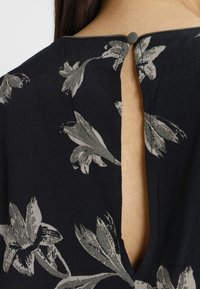 Svart blommig topp med en ryggdesign i form av ett nyckelhål, med grå blomstermönster och en knappstängning vid kragen. Slät tygtextur.