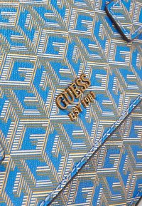 Blå og guld mønstret stof med gentagne geometriske former, der har et guld "GUESS EST 1981" logo på syet materiale.