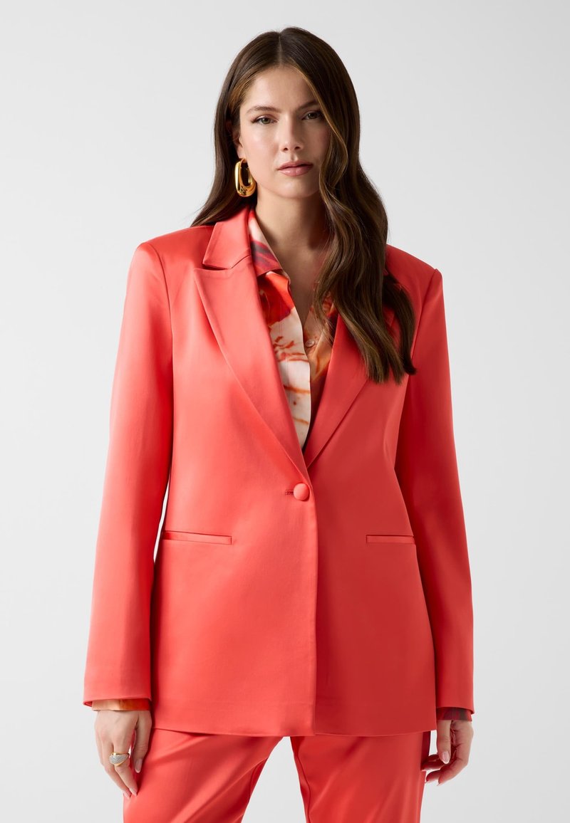 Femme aux longs cheveux bruns portant un blazer orange et un pantalon assorti par-dessus une chemise à motifs, debout devant un fond gris clair.