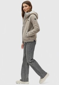 Hoodie en polaire beige avec une texture pelucheuse, capuche à cordon de serrage et doublure claire contrastante ; associé à un jean droit gris et des sneakers claires.