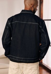 Giacca in denim scuro con cuciture a contrasto, colletto standard e tasche laterali, indossata sopra pantaloni beige, mostrante la vista posteriore.