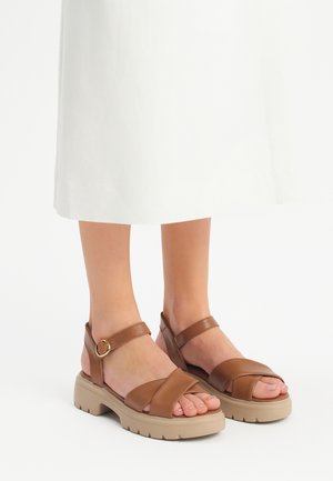 Brown leather sandals with crisscrossed straps and ankle buckle. Thick beige outsole with a textured grip. Toes visible.