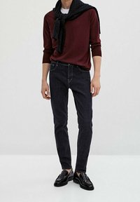 Donkerdenim jeans gekleed met een bordeauxrode lange mouw shirt, voorzien van een zwarte gebreide sjaal over de schouders gedrapeerd en zwarte instappers.