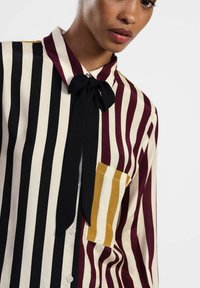 Donna che indossa una camicia a righe con strisce nere, crema e bordeaux, un papillon nero e una tasca a righe giallo senape sul petto.