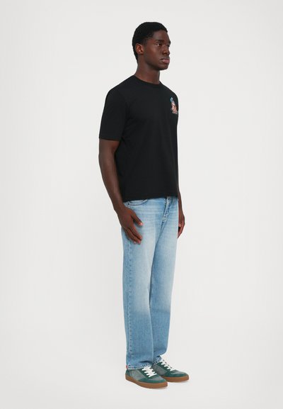 Man standing in light blue jeans, black graphic t-shirt, and green sneakers against plain white background.