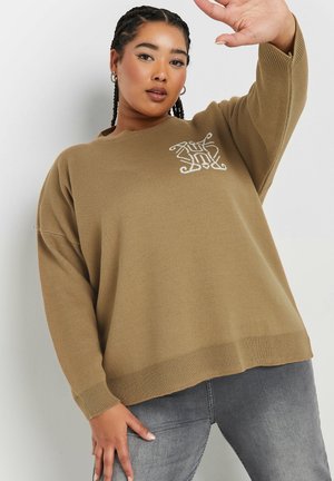 Pull en tricot marron avec un col rond, des épaules tombantes et un motif brodé sur la poitrine, associé à un pantalon gris.