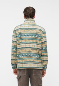 Άνδρας που φοράει φλις σακάκι με μοτίβο σε οριζόντιες ρίγες μπλε, μπεζ και κρεμ, γυρισμένος ανάποδα, σε συνδυασμό με καφέ παντελόνι cargo.