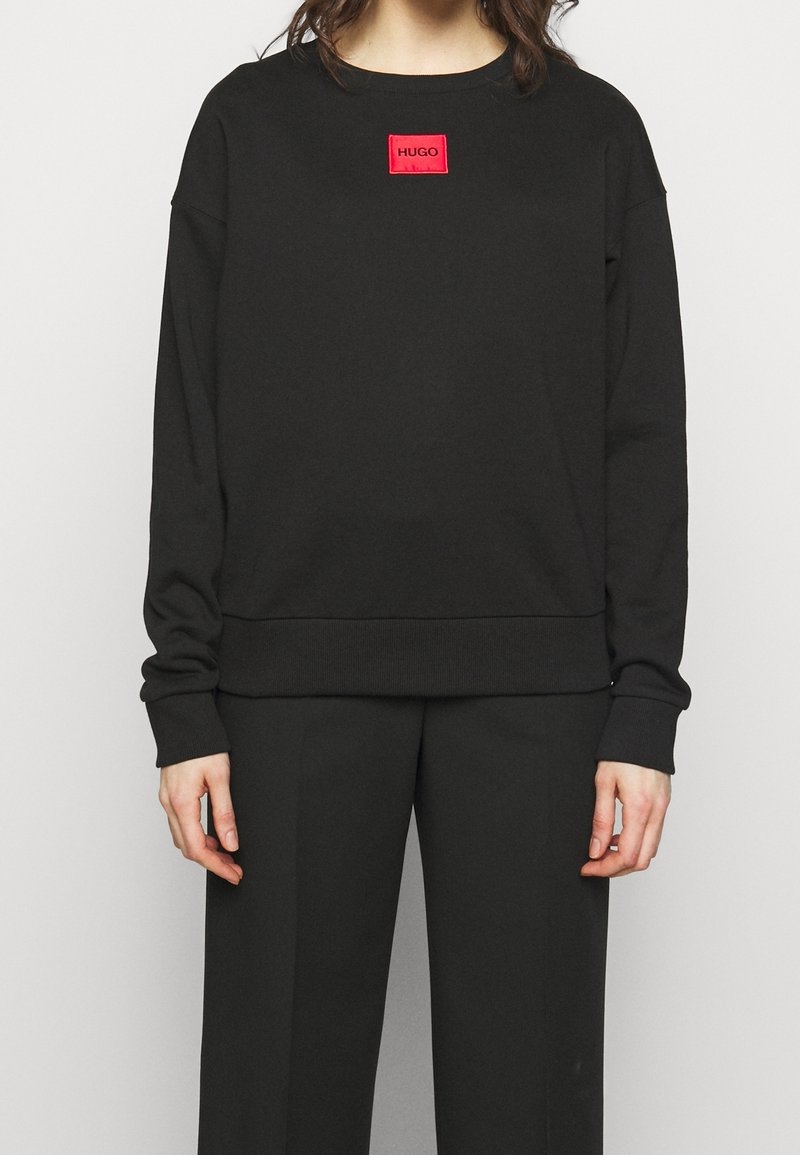 Sudadera negra con mangas largas, puños y dobladillo acanalados, con una etiqueta cuadrada roja con el texto "HUGO" en el frente. Combinada con pantalones negros.