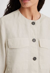 Lys beige linned skjorte med rund halsudskæring, tre sorte knapper og to brystlommer med klapper. Tekstureret stof med en naturlig finish.