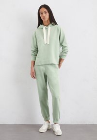 Lichtgroene hoodie met witte trekkoorden, gecombineerd met bijpassende broek. De stof lijkt zacht, met een ontspannen pasvorm en geribde boorden. Gestyled met lichte sneakers.