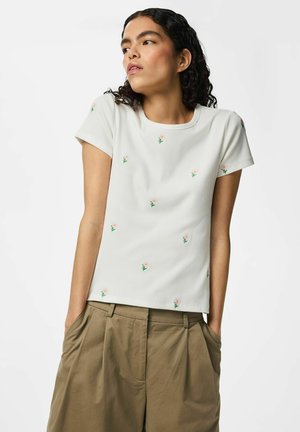 Jeune femme aux cheveux foncés bouclés portant un haut blanc à manches courtes avec de petits motifs floraux et un pantalon kaki ample, les mains dans les poches.