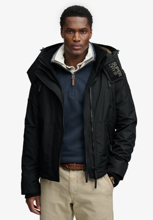 Mężczyzna w czarnej kurtce Superdry na granatowym swetrze rozpinanym do połowy i beżowych spodniach, stoi z jedną ręką w kieszeni na białym tle.