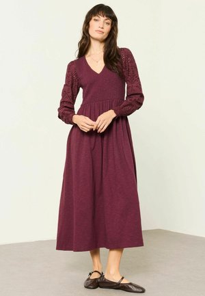 Donna con un lungo vestito color porpora con maniche a sbuffo strutturate e scarpe basse nere, in piedi su uno sfondo chiaro e semplice.