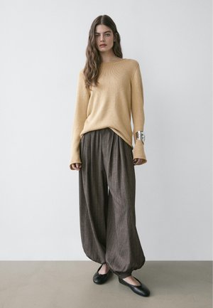 Frau mit langem braunem Haar, die einen beigen Pullover, weite braune Hose mit Nadelstreifen und elastischen Säumen sowie schwarze flache Schuhe trägt und vor einer schlichten Wand steht.