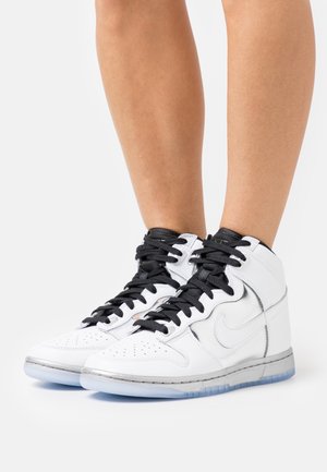 Baskets montantes Nike blanches avec des lacets noirs portées sur des jambes nues, debout sur une surface blanche.