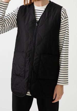 Osoba nosząca czarną pikowaną kamizelkę bez rękawów z przednimi kieszeniami na białej i czarnej pasiastym długim rękawem.