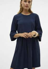 Marinblå knälång klänning med trekvartsärmar, mjuk textur, rund halsringning och en lös passform. Modellen bär ett ljust armband.